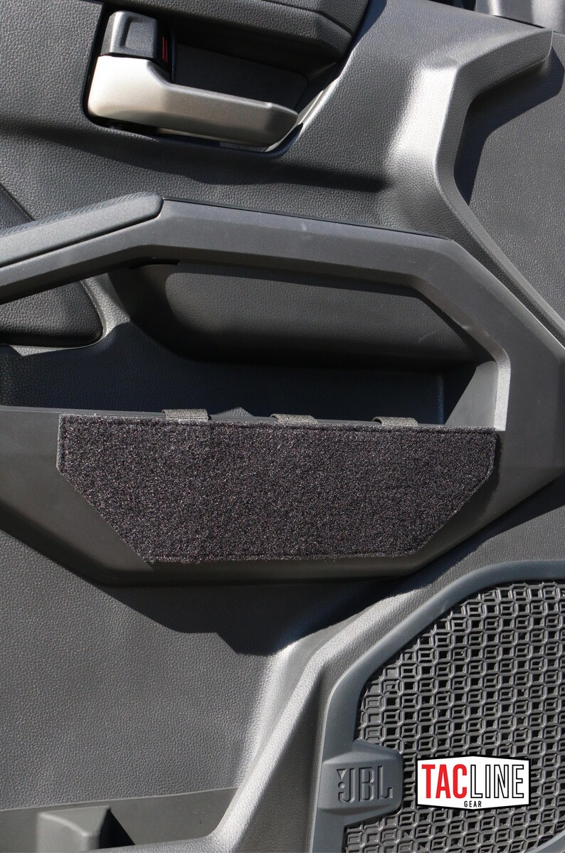 May include: Black interior door panel with a textured surface. A black, rectangular, fabric-covered insert is placed in the door handle area. The JBL and TACLINE GEAR logos are visible.