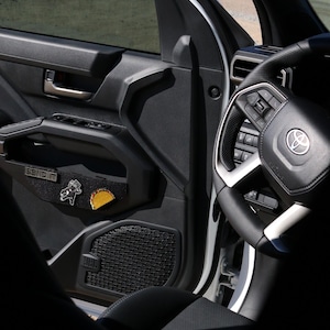 May include: Interior view of a vehicle's front door, featuring a black door panel with a "SEND IT" patch and two decorative pins. The steering wheel and part of the dashboard are visible, with a scenic background through the window.