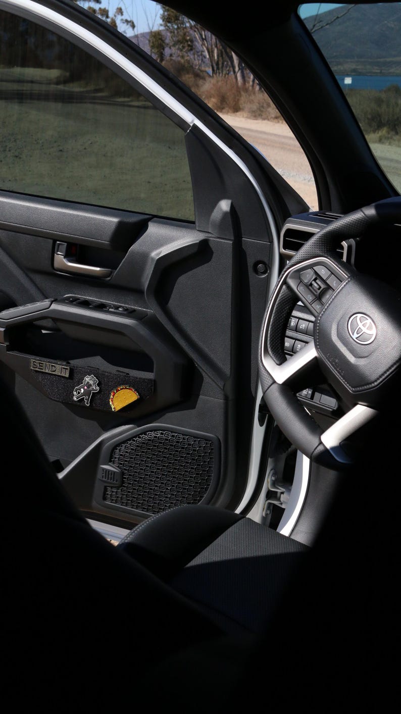 May include: Interior view of a vehicle's front door, featuring a black door panel with a "SEND IT" patch and two decorative pins. The steering wheel and part of the dashboard are visible, with a scenic background through the window.