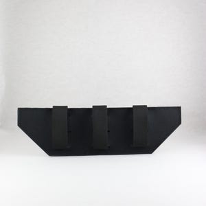 May include: A black, angular pouch with three vertical straps. The pouch has a trapezoidal shape with a flat top and angled sides. The straps are evenly spaced across the top of the pouch.