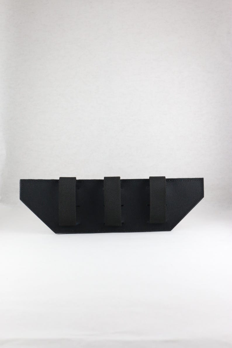 May include: A black, angular pouch with three vertical straps. The pouch has a trapezoidal shape with a flat top and angled sides. The straps are evenly spaced across the top of the pouch.