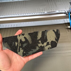 May include: A camouflage-patterned tactical gear accessory, held in a hand. The accessory features a mix of black, gray, and olive green colors. The item is shown against a backdrop of industrial equipment.