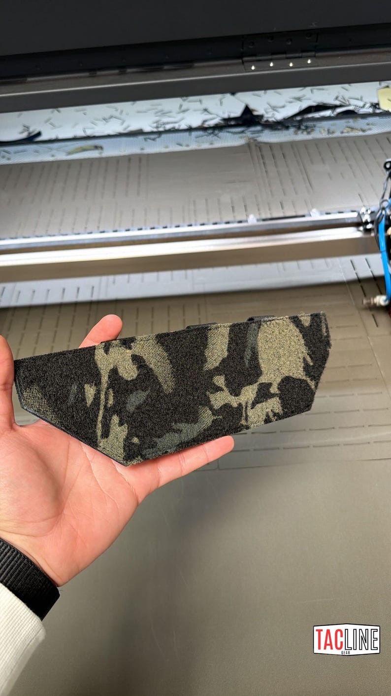 May include: A camouflage-patterned tactical gear accessory, held in a hand. The accessory features a mix of black, gray, and olive green colors. The item is shown against a backdrop of industrial equipment.