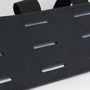 May include: Close-up of a black tactical gear component. The fabric has multiple horizontal slits and two attached loops. The item is made of a durable, textured material, and is designed for attaching additional accessories.