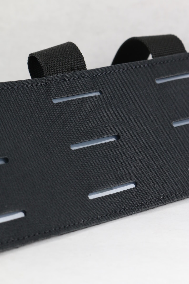 May include: Close-up of a black tactical gear component. The fabric has multiple horizontal slits and two attached loops. The item is made of a durable, textured material, and is designed for attaching additional accessories.