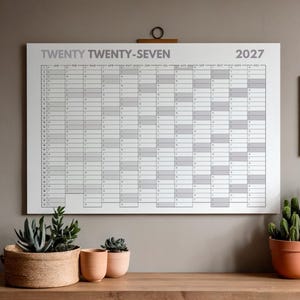 Może przedstawiać: Duży biały planer roczny na rok 2027 z szarymi liniami siatki i napisem "TWENTY TWENTY-SEVEN" wisi na neutralnej ścianie. Drewniany stół poniżej prezentuje rośliny doniczkowe i oprawione czarno-białe zdjęcie krajobrazu.