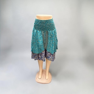 May include: A teal and patterned skirt with a smocked waistband. The skirt has a layered design, with a teal floral print over a patterned, dark blue and purple layer. The skirt is displayed on a mannequin.