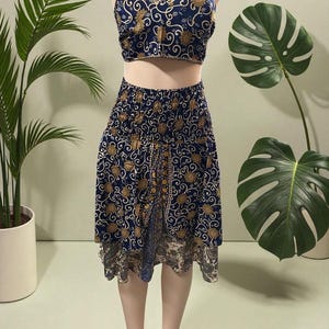 May include: A two-piece halter top and skirt set on a mannequin. The navy blue fabric is patterned with gold floral and swirl designs. The skirt is knee-length with a split front. The top has a V-neck and a halter tie.