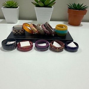 May include: A collection of colorful bracelets and small bowls. The bracelets are made of various materials and feature different patterns and colors, including orange, floral, and brown. The bowls are black, red, purple, brown, and blue. Green succulents in white and terracotta pots are in the background.
