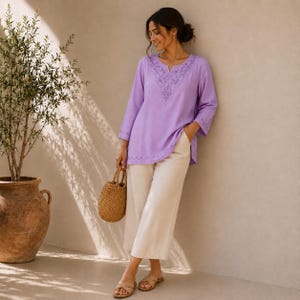 May include: A light purple long-sleeved tunic featuring a V-neckline with intricate tonal embroidery, paired with loose-fitting beige trousers. The tunic has a scalloped hemline detail and is worn with tan slide sandals and a woven straw handbag.
