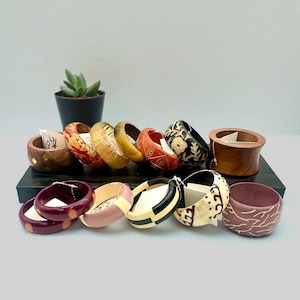 May include: A collection of wooden bangles in various colors and designs. The bangles are displayed on a black surface, with a small potted plant in the background. The bangles feature carved patterns, painted details, and a range of colors including red, yellow, and black.