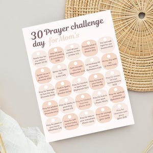 Puede incluir: Un desafío de oración impreso para mamás, con 30 indicaciones diarias con versículos bíblicos. El diseño incluye una combinación de colores rosa claro y blanco. La hoja se coloca sobre una superficie neutra con una cesta tejida y flores secas.