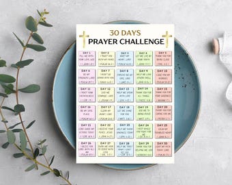 May include: A printed 30 Days Prayer Challenge card with a gold cross design, displayed on a light blue plate. The card features a grid of colorful squares with daily prayer prompts. A sprig of greenery and a roll of white lace ribbon are also present.
