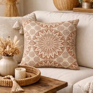 May include: A decorative throw pillow with a beige background and a repeating geometric pattern in shades of brown. The pillow is square and rests on a neutral-colored sofa. The design has a floral or mandala-like appearance.