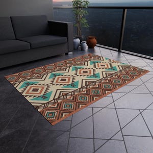 May include: Rectangular outdoor rug with a geometric pattern in shades of brown, teal, and cream. The rug features a border with diamond shapes and a central design of triangles and squares. The rug is placed on a tiled surface.
