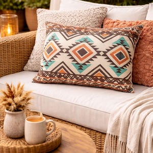 May include: A decorative pillow with a geometric pattern in brown, orange, and teal on a cream background. The pillow is on a white sofa with other pillows and a blanket. A wooden table holds a tray with mugs and a vase.