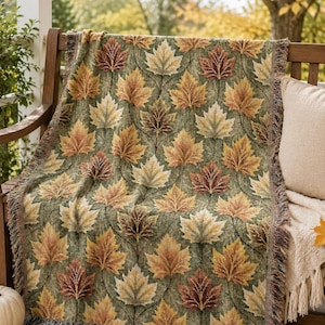 May include: A woven throw blanket featuring a repeating pattern of maple leaves in shades of orange, yellow, and brown against a muted green background. The blanket has a textured finish with fringed edges along the sides and bottom hem.