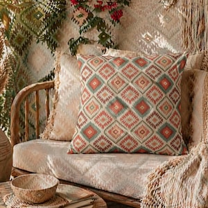 Op de afbeelding: Een decoratief sierkussen met een geometrisch ruitpatroon in rood, groen en beige tinten. Het kussen ligt op een beige bank, samen met andere kussens en een geweven deken. De scène roept een gezellige, bohemien sfeer op.