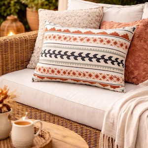 May include: A decorative pillow with a geometric pattern in shades of orange, black, and gray rests on a white couch. Other pillows in neutral tones and a fringed throw blanket complete the cozy outdoor seating arrangement.