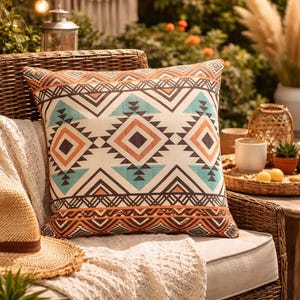 May include: A decorative throw pillow with a geometric pattern in shades of orange, teal, and brown. The pillow is on a wicker chair with a cream-colored blanket and a straw hat. The outdoor setting includes plants and string lights.