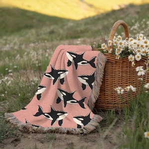 May include: A pink throw blanket with a repeating pattern of black and white orca whales. The blanket has fringed edges and is draped over a picnic basket filled with white daisies. The scene is set outdoors in a field.