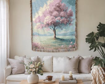 May include: A woven tapestry depicting a pastel landscape with a large pink flowering tree. The artwork has a fringed border and hangs above a white sofa with neutral-toned cushions. A wooden coffee table displays a vase of flowers and candles.