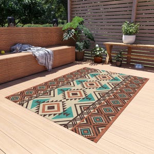 May include: Rectangular outdoor rug with a geometric pattern in shades of brown, teal, and cream. The rug features diamond and triangle shapes, creating a Southwestern or tribal design. It is placed on a wooden deck, suggesting an outdoor living space.