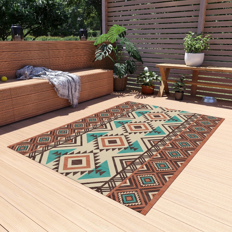 May include: Rectangular outdoor rug with a geometric pattern in shades of brown, teal, and cream. The rug features diamond and triangle shapes, creating a Southwestern or tribal design. It is placed on a wooden deck, suggesting an outdoor living space.