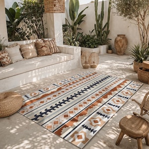 May include: A rectangular outdoor rug with a geometric pattern in shades of cream, brown, navy, and gray. The rug is placed on a concrete patio, with a seating area and plants in the background. The rug's design features stripes and diamond shapes.