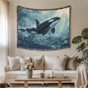 Puede incluir: Un tapiz tejido con una ilustración detallada de una orca nadando bajo el agua. La obra de arte muestra las marcas blancas y negras de la ballena sobre un fondo de tonos azules y verdes, con un borde con flecos.