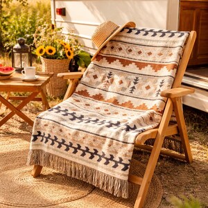 May include: A wooden folding chair with a patterned blanket and straw hat sits outside a camper. The blanket features geometric designs in brown, beige, and navy blue. A round jute rug is beneath the chair. A small table with a cup and watermelon is nearby.