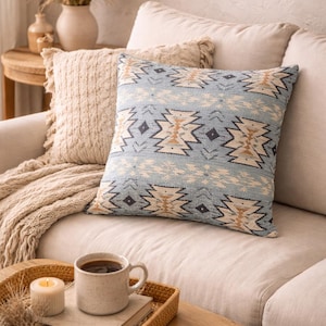 May include: A light blue decorative pillow with a geometric pattern rests on a beige sofa. A woven tray holds a coffee mug, a candle, and a book. The scene evokes a cozy, bohemian living room aesthetic.