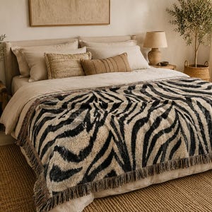 May include: A rectangular throw blanket featuring a black and beige zebra stripe pattern with a fringed edge. The blanket is draped over a bed with neutral-colored bedding, including several pillows in shades of cream, tan, and brown.