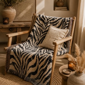 May include: A light-colored wooden armchair draped with a woven throw blanket featuring a black and beige zebra stripe pattern and fringed edges. A square, textured beige throw pillow rests on the seat of the chair against the backrest.