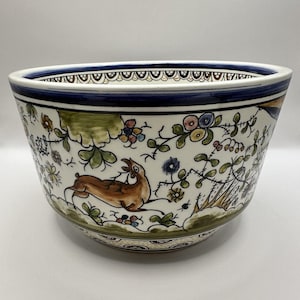 May include: A ceramic bowl with a white base featuring hand-painted illustrations of a brown deer, green foliage, and colorful flowers. The rim is decorated with a dark blue band and a scalloped pattern, creating a detailed, multi-colored design.
