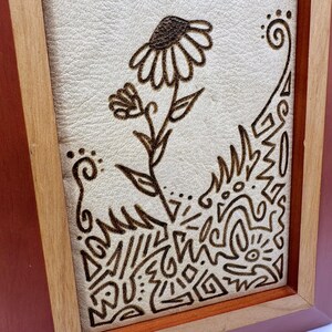 May include: Wooden framed artwork featuring a detailed pyrography design. The artwork showcases a stylized flower with intricate patterns and shapes. The frame is a warm brown color, and the background is a light beige.