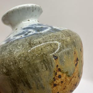 May include: A small ceramic vase with a rounded body and a flared rim. The surface features a mottled glaze with earthy tones of olive green, brown, and tan, transitioning to a white speckled glaze around the neck and rim with blue accents.