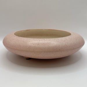 May include: A low-profile, circular ceramic bowl with a speckled light pink exterior and a smooth, cream-colored interior. The vessel features a wide, rounded rim and is supported by small, discreet feet, presenting a simple, functional design.