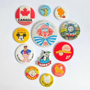 May include: A selection of vintage pin badges with diverse designs. Includes a red Canada badge, a Mickey Mouse badge, a clown badge, and a Thomas the Tank Engine badge. Other badges feature cartoon characters and text.