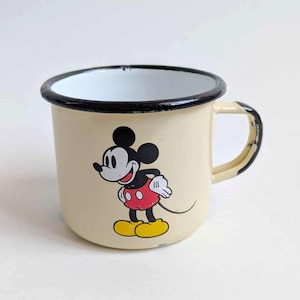 May include: Cream-colored enamel mug featuring a classic Mickey Mouse design. The mug has a black rim and handle, with Mickey in his iconic red shorts, yellow shoes, and white gloves. A vintage-style collectible.