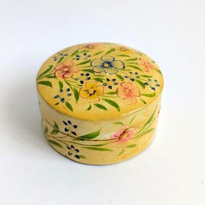 May include: A round, yellow lidded box featuring hand-painted floral motifs in shades of pink, blue, and yellow, accented with green leaves. The cylindrical container has a smooth, glossy finish and a matching lid that fits securely on top.
