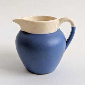 Könnte beinhalten: Ein kleiner, blauer Keramikkrug mit cremefarbenem Rand und Griff. Der Krug hat einen abgerundeten Körper und einen Ausguss zum Ausgießen. Der Griff ist ebenfalls am Boden blau. Der Krug wird wahrscheinlich zum Servieren von Flüssigkeiten verwendet.