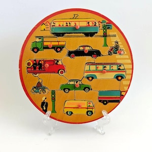 May include: A vintage wooden puzzle featuring various vehicles, including a bus, car, and truck, arranged in a circular pattern. The puzzle has a yellow background with red and green accents, and is mounted on a clear stand.