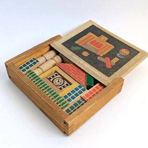 May include: A wooden box containing various building blocks with painted patterns including a clock face, red brick designs, and blue grid motifs. The sliding lid features a black panel with a printed illustration of a building structure.