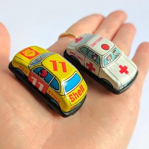 Peut inclure: Deux petites voitures anciennes : une voiture de course jaune avec le numéro 11 et la marque Shell, et une ambulance blanche avec des croix rouges. La voiture de course a des fenêtres bleues et l'ambulance a un feu rouge sur le dessus.