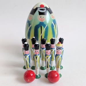 Peut inclure: Ensemble de soldats jouets en bois peints, vêtus d'uniformes jaunes, bleus et blancs, devant une figurine décorative en forme d'œuf. Deux boules rouges en bois sont au premier plan. Les jouets ont des chapeaux noirs.