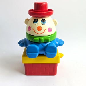 Könnte beinhalten: Ein Vintage-Spielzeugclown aus Kunststoff mit rotem Hut, blauen Armen und Beinen und einer grünen Fliege. Der Clown hat ein lächelndes Gesicht mit rosigen Wangen und sitzt auf einem gelb-roten Block.