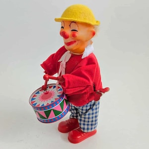 Puede incluir: Un juguete de payaso de cuerda vintage. El payaso lleva un sombrero amarillo, una chaqueta roja y pantalones a cuadros azules y blancos. Sostiene un tambor colorido con un diseño rosa y verde y baquetas rojas. Los zapatos rojos completan el atuendo.