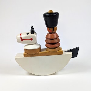 May include: A wooden toy featuring a white rocking base with a black tail, topped with a white horse head with red bridle details and a figure wearing a tall black hat, brown layered clothing, and a painted face, all mounted on a flat wooden plank.