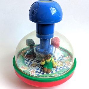 Puede incluir: Un juguete de peonza con una cúpula de plástico transparente que alberga tres pequeñas figuras sobre una base de picnic. El juguete tiene un mango azul, un borde verde y una base roja. La base muestra un mantel de picnic a cuadros azules.